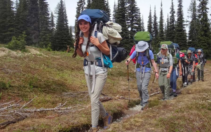 A group of people  wearing backpacks hike along a trail. There is a line of evergreen trees in the background.